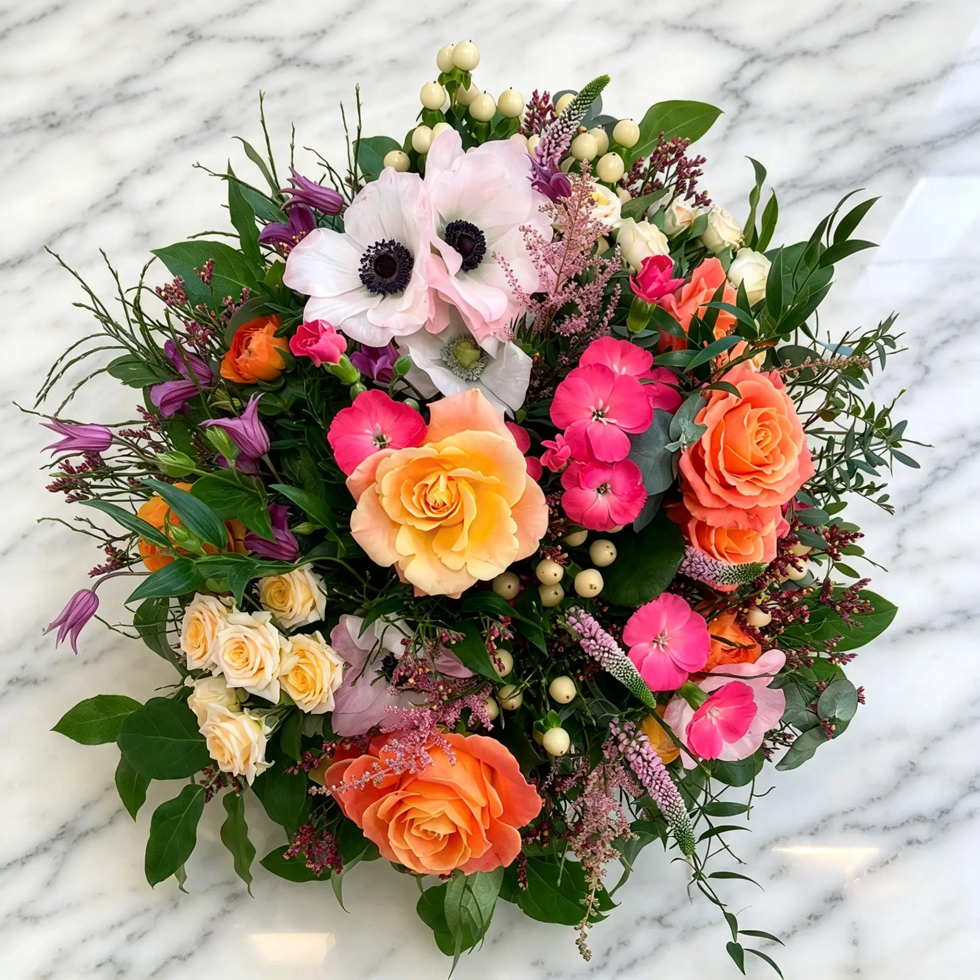 Rund gebundener bunter Strauß mit Anemonen, orangefarbenen Rosen und rosa Blüten auf Marmor