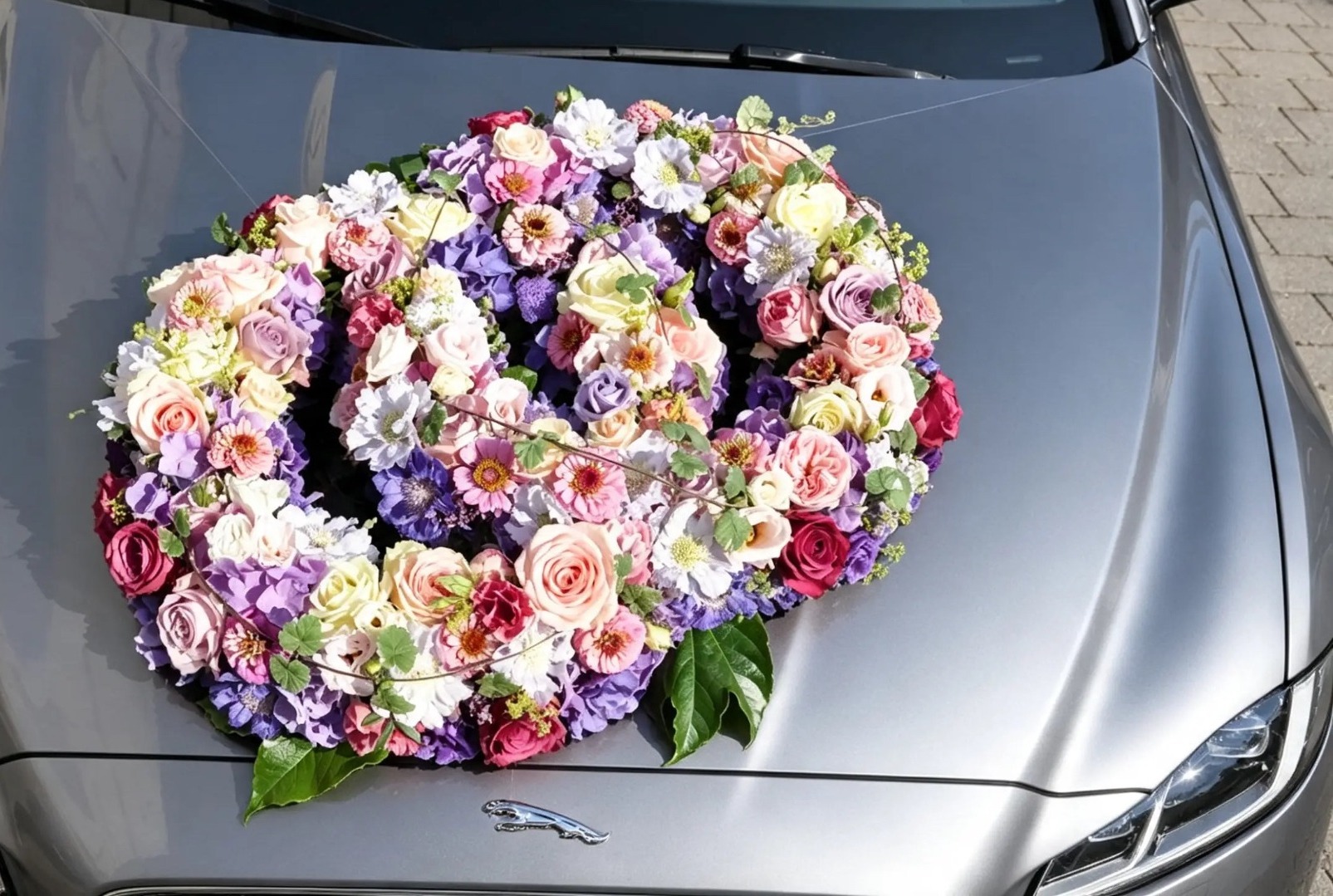 Großes Blumenherz aus Rosen, Hortensien und feinen Blüten auf einer grauen Motorhaube
